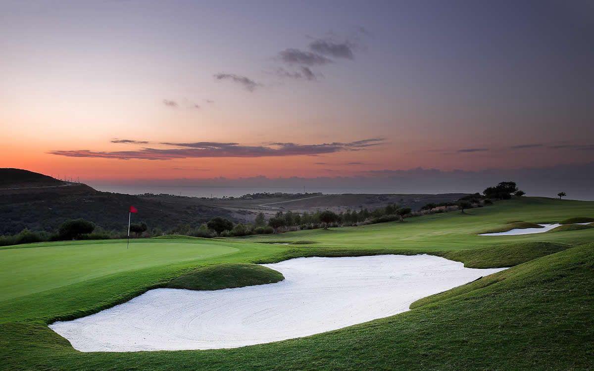 A manicured green next to a sand bunker with the sun setting in the distance
