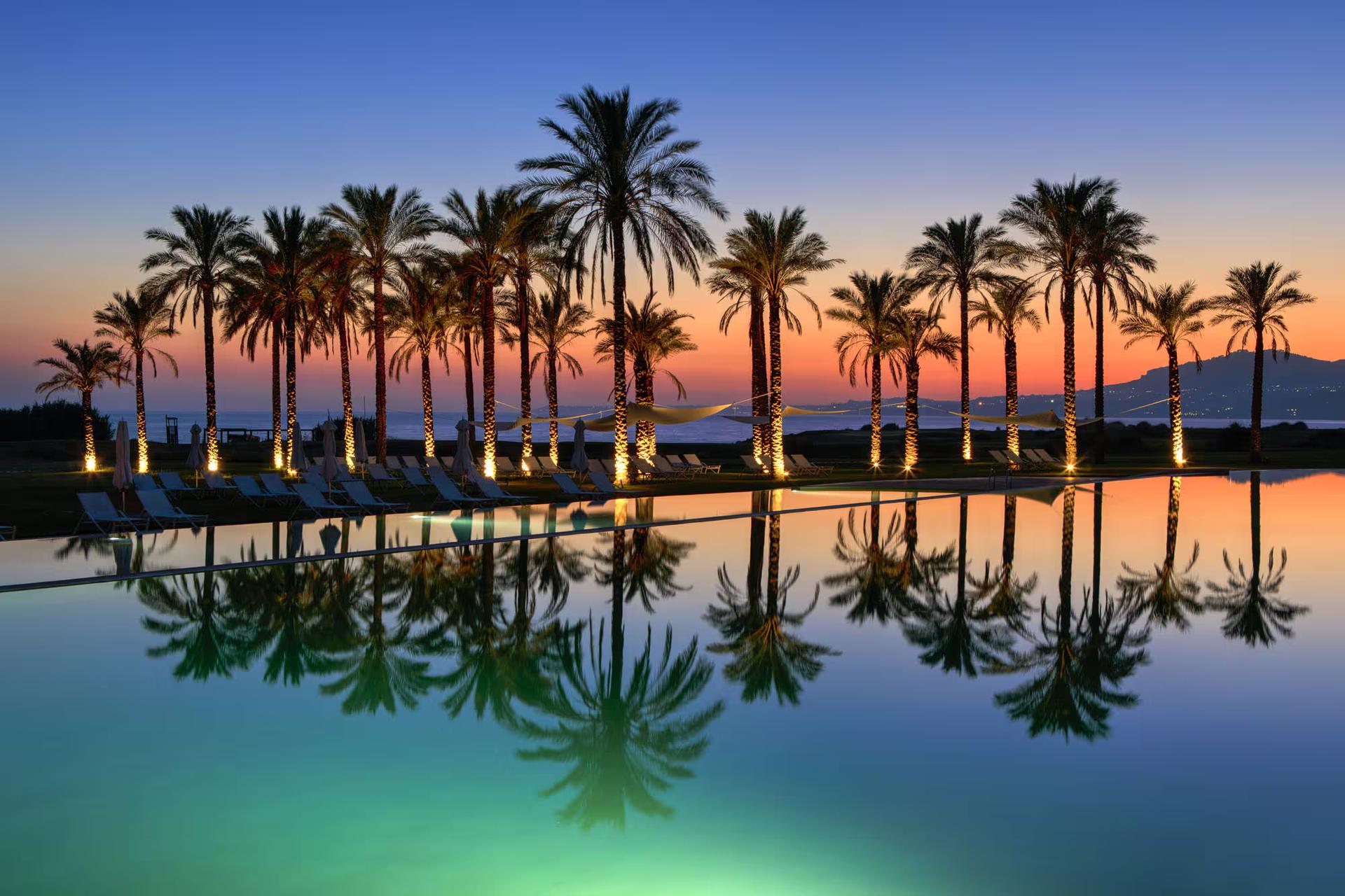 Outdoor swimming pool at Verdura Resort, Rocco Forte Hotels surrounded by palm trees at sunset